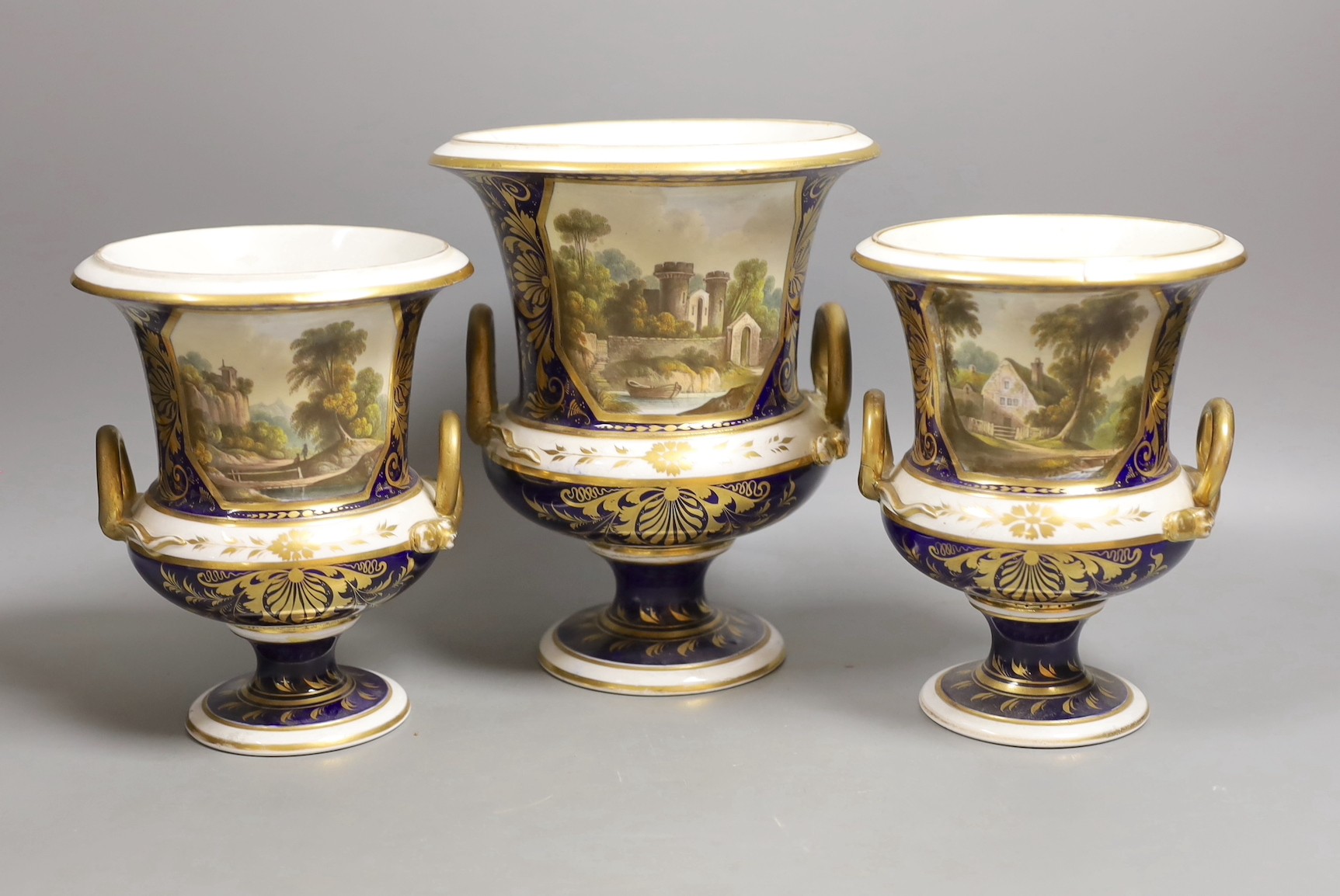 A garniture of three Derby named twin handled cups, View near Belper, Derbyshire, and 'View in North Wales' and 'View near Rome', 17cm and 20.5cm